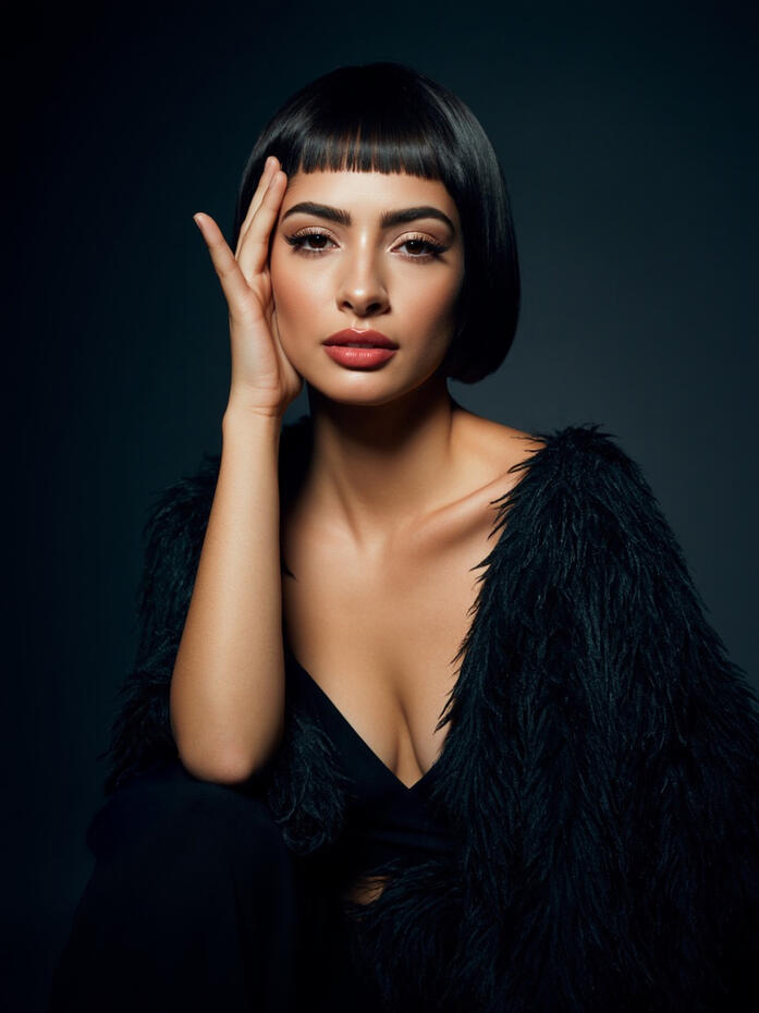 Baseerat Al-Rumayyan in a black textured coat, seated against a dark backdrop with hand resting on face, wearing a short modern bob haircut with fringe—studio lighting highlighting her luminous makeup and confident expression.